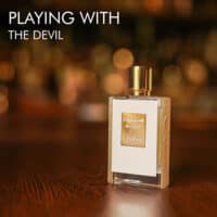 A 50ml bottle of Kilian Playing With The Devil Eau de Parfum with a white glass body, gold-tone accents, and a rectangular shape, set on a brown table.