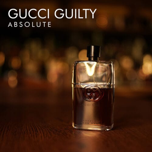 a 90ml bottle of Gucci Guilty Absolute Eau de Parfum with a square glass design featuring a deep amber liquid, a gold Gucci logo, and a black cap on a brown table.