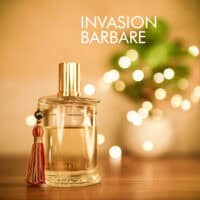 A 100ml bottle of Parfums MDCI Invasion Barbare Eau de Parfum with a rounded glass design and a white porcelain bust-shaped cap, placed on a brown table.