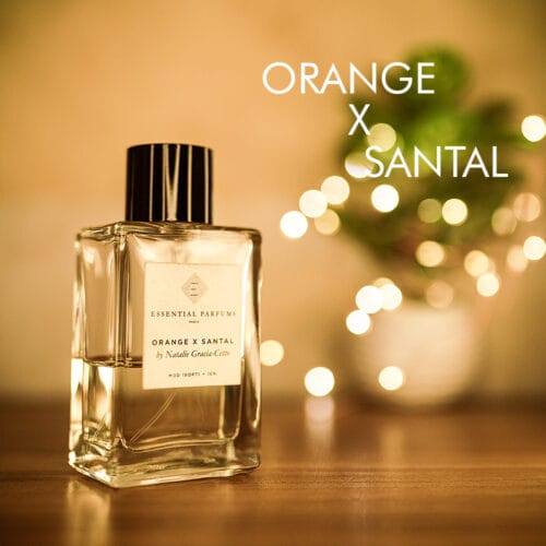 A 100ml bottle of Essential Parfums Orange x Santal Eau de Parfum with a square glass bottle, transparent with a white label, and a chrome cap, placed on a brown table.