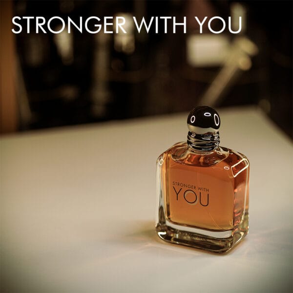 A 100ml bottle of Giorgio Armani Stronger With You Eau de Toilette with a clear glass bottle, dark brown accents, and a silver-toned cap, displayed on a white table.