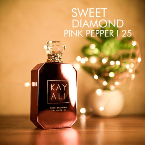 A 100ml bottle of Kayali Sweet Diamond Pink Pepper | 25 Eau de Parfum with a clear cap and a pink shade glass body, set on a brown table.