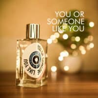 A 100ml bottle of Etat Libre D’Orange You Or Someone Like You Eau de Parfum with a tall, clear glass bottle, featuring a white label with black text, and a silver cap, placed on a brown table.