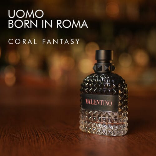 a 100ml bottle of Valentino Uomo Born In Roma Coral Fantasy Eau de Parfum with an orange studded glass design and black cap on a brown table