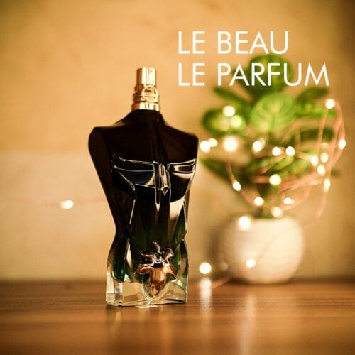 a 125ml bottle of Jean Paul Gaultier Le Beau Le Parfum Eau de Parfum Intense with a torso-shaped dark green glass design featuring a gold leaf detail at the waist and a gold cap on a brown table.