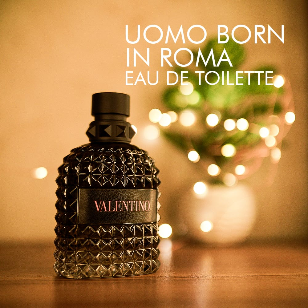 a 100ml bottle of Valentino Uomo Born in Roma Eau de Toilette with a transparent glass design, black studs, and a black cap on a brown table.