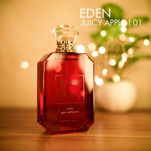 A 100ml bottle of Kayali Eden Juicy Apple | 01 Eau de Parfum with a red glass body, gold label, and a clear cap on a brown table.