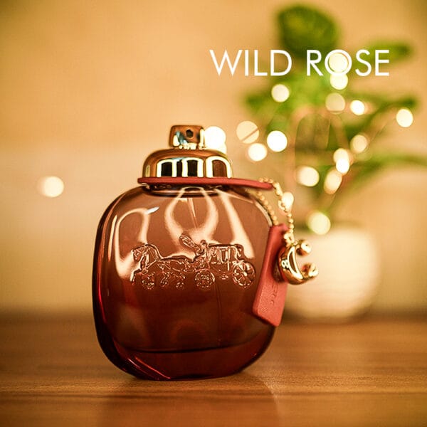 A 90ml bottle of Coach Wild Rose Eau de Parfum with a pink glass body, a gold cap, and a gold charm attached to the neck on a brown table.