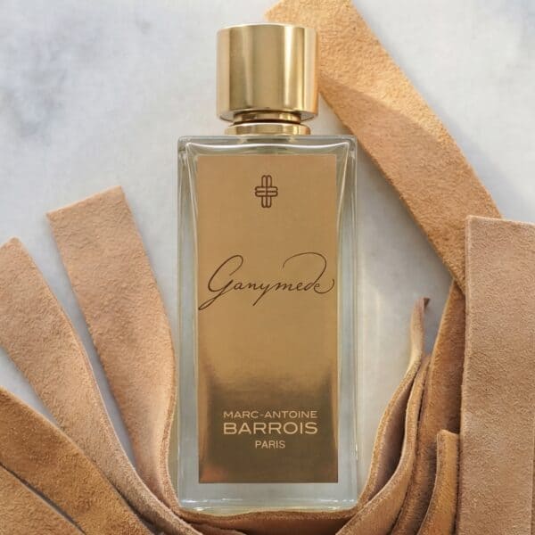 1 Elegant Marc-Antoine Barrois Ganymede perfume bottle with a beige label and gold cap, surrounded by soft beige leather strips.